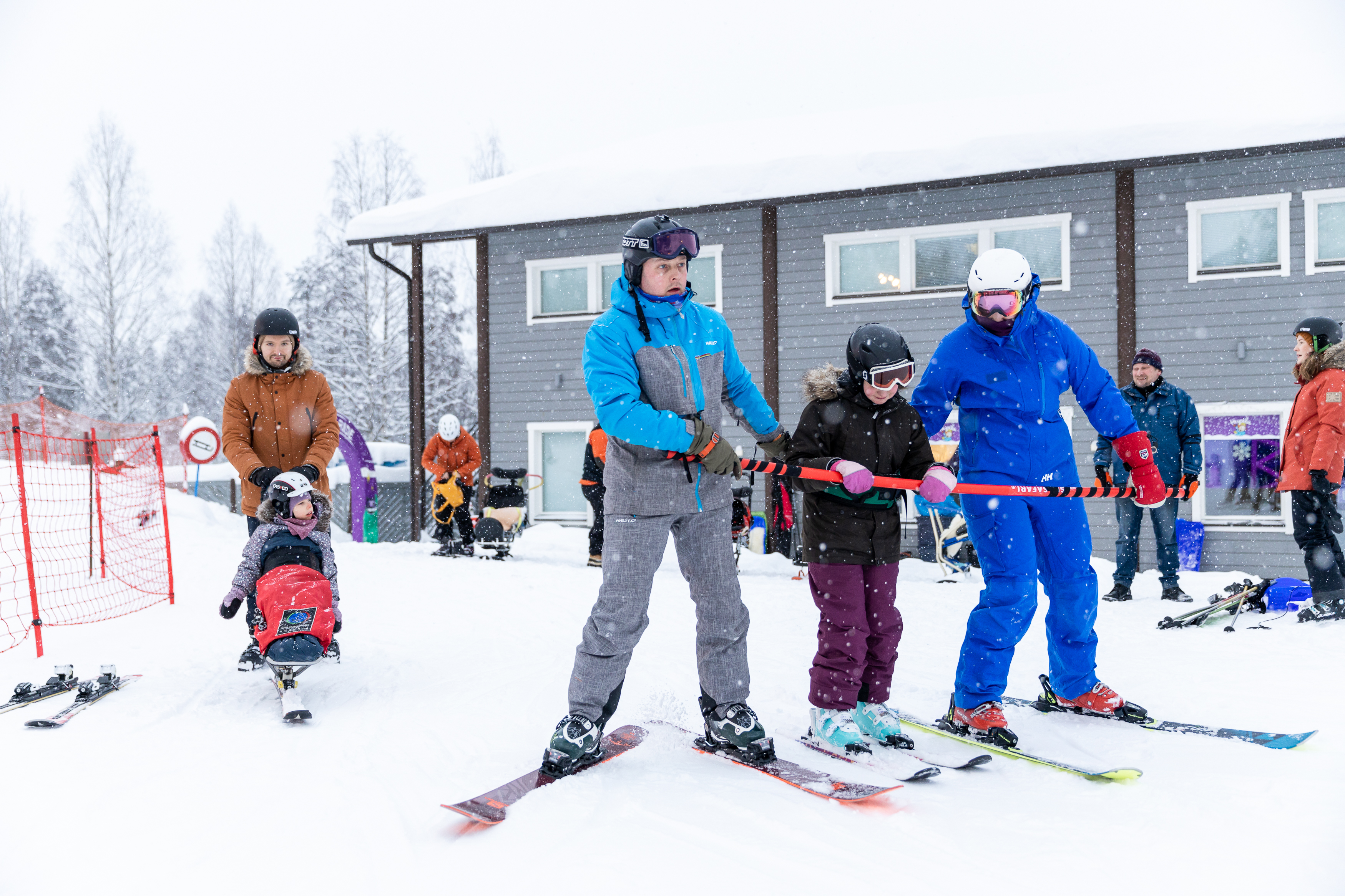 Kuvassa kaksi henkilöä suksilla avustaa henkilöä punaisen sauvan avulla.